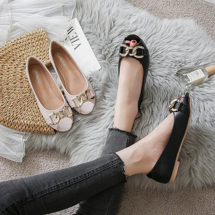 A pair of black peep-toe flat shoes with gold chain detailing on the vamp, worn by a person in dark jeans on a grey rug, alongside a pair of cream flats.
