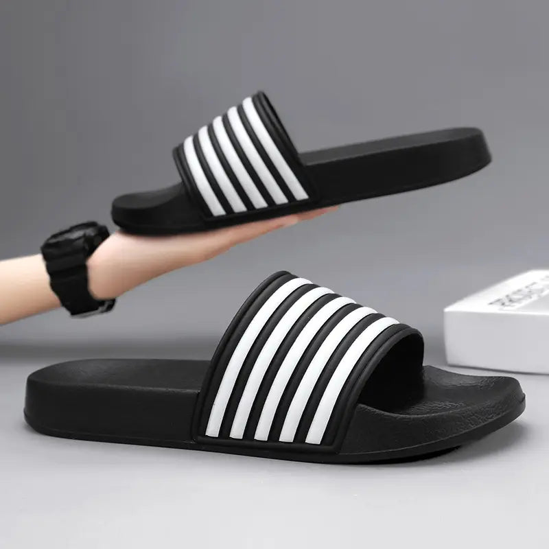A pair of black Zylos EVA comfort slides with iconic white stripe detailing on the strap, presented on a studio background. One slide is held in a hand, showcasing the design.