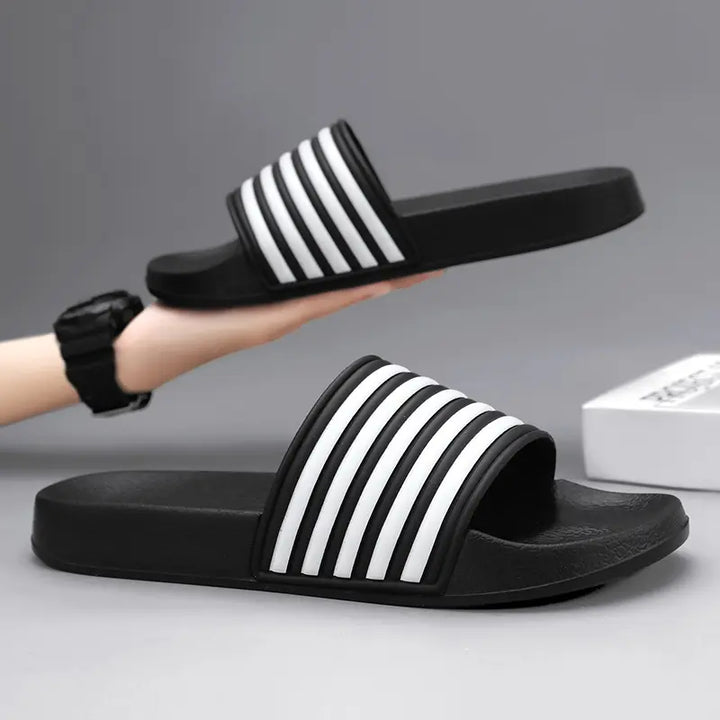 A pair of black Zylos EVA comfort slides with iconic white stripe detailing on the strap, presented on a studio background. One slide is held in a hand, showcasing the design.