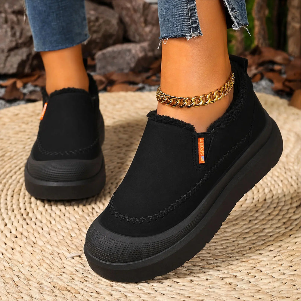 Black Zylos slip-on comfort boots with plush shearling lining and a chunky grey platform sole, styled on feet with cuffed jeans and a gold anklet.