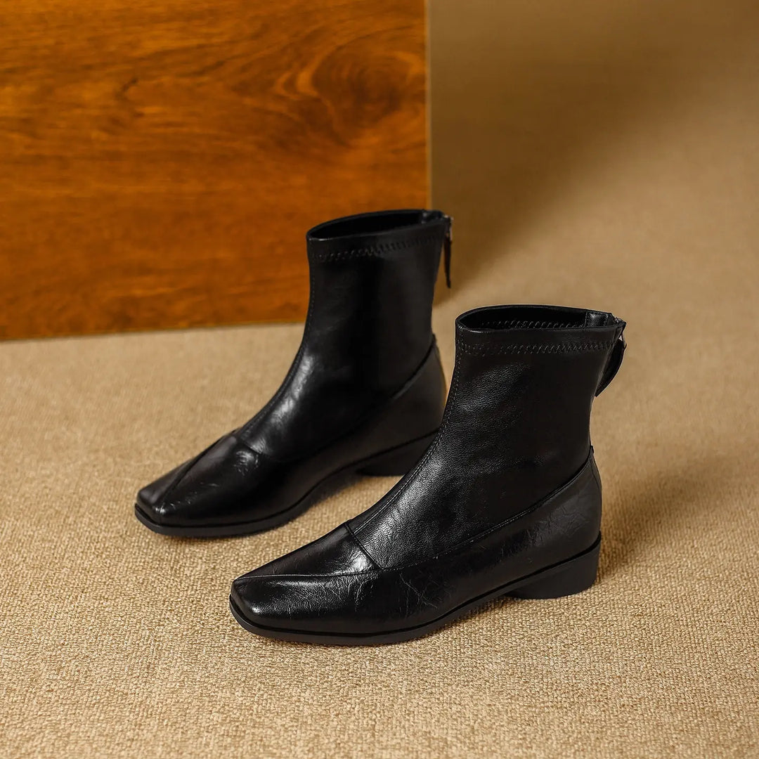 Zylos black leather-look ankle boots with a contemporary square toe and a low block heel, shown from a three-quarter angle on a textured studio background.