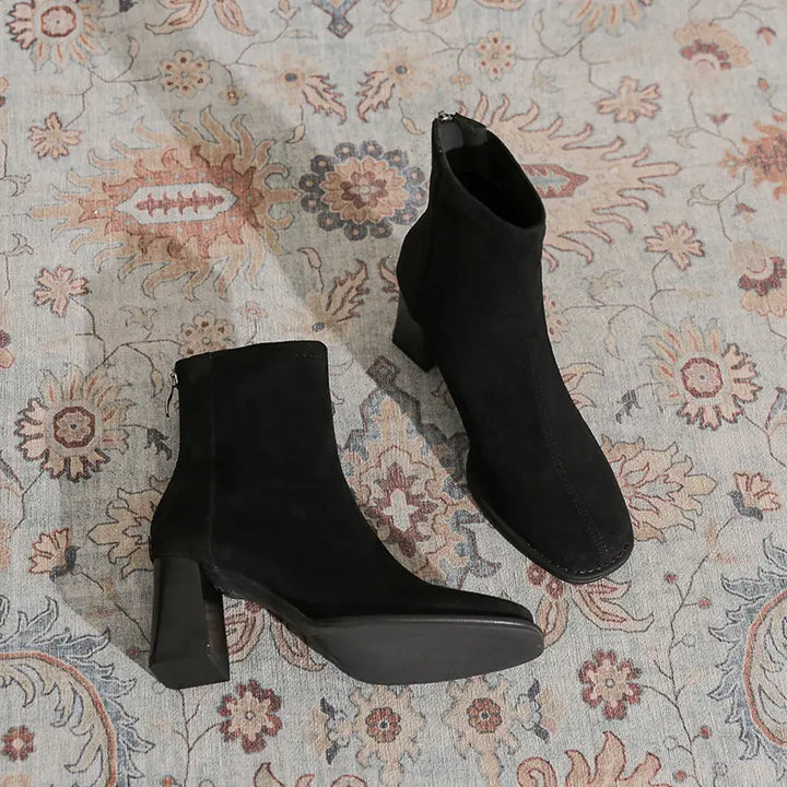 Zylos black suede-like ankle boots with square toe, chunky block heel, and back zipper, artfully arranged on a decorative rug. These chic winter boots offer timeless style and sophisticated comfort.