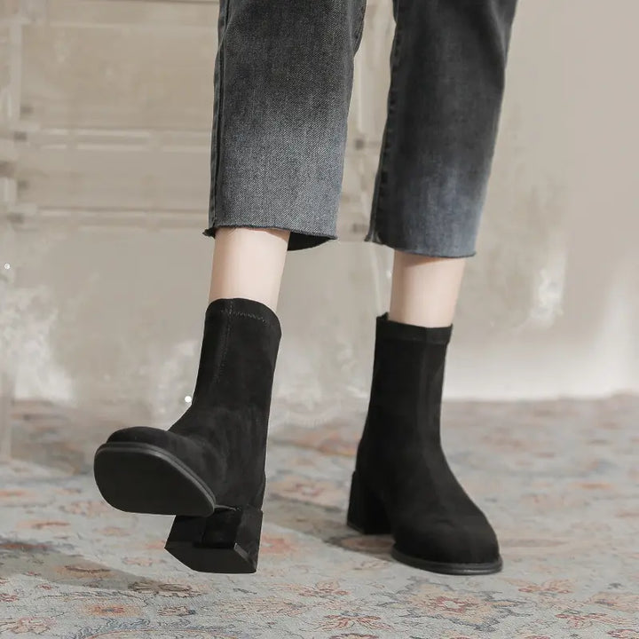 Black suede-like Zylos ankle boots with square toes and block heels, worn with cropped jeans by a model, showcasing a chic winter footwear style.