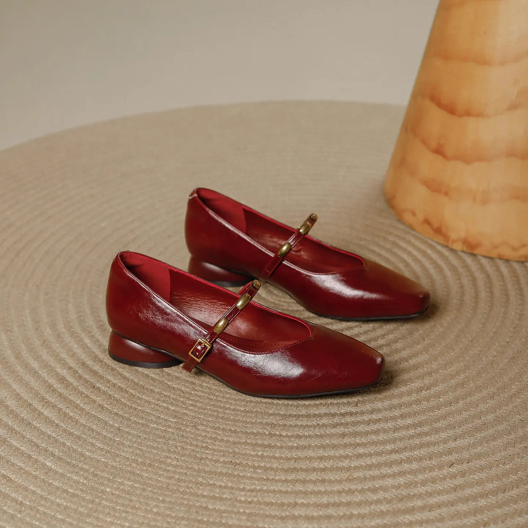 Pair of burgundy wine Zylos square toe Mary Jane shoes with low block heels and gold stud buckle straps, shown on a textured rug.