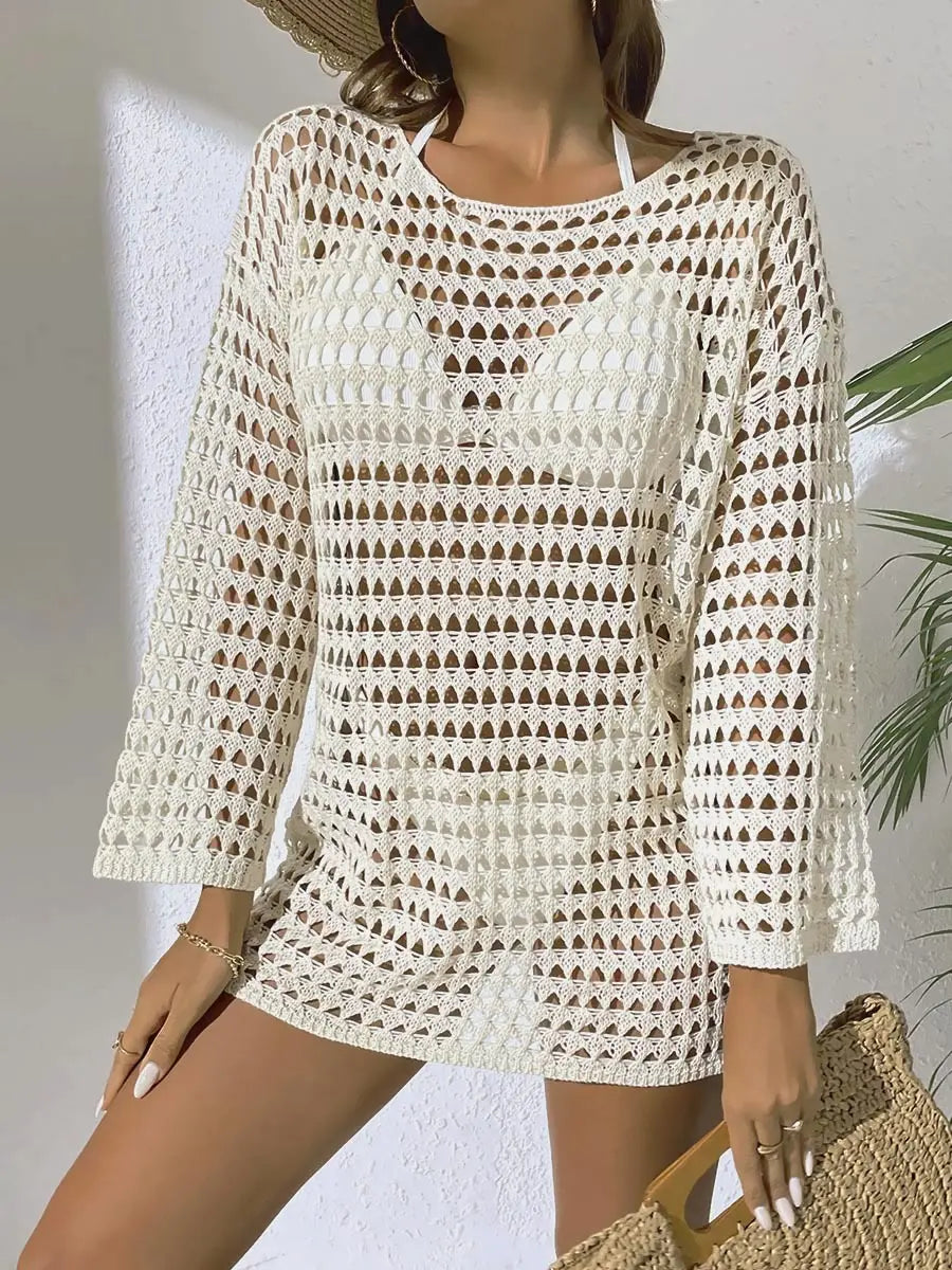 Cream crochet beach cover-up dress worn by a model over white swimwear, featuring an intricate open-knit pattern, round neckline, and long sleeves in a sunny resort setting.