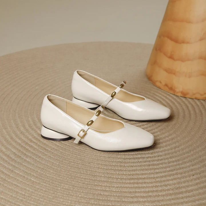 Ivory cream square-toe Mary Jane shoes with a low block heel, featuring a slim strap adorned with gold-tone studs and buckle, presented on a textured rug.
