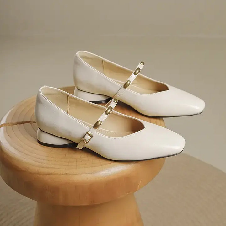 Two cream-colored Zylos Square Toe Mary Jane shoes with low block heels and gold-tone stud-embellished straps are displayed at a three-quarter angle on a wooden stool.