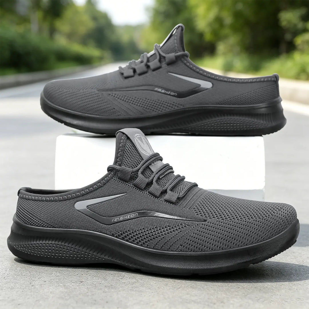 A pair of dark grey Zylos breathable comfort slip-on trainers shown from a three-quarter angle, with a second pair slightly behind. The shoes feature engineered mesh uppers, elastic laces, and a matte black sole with a reflective side stripe.