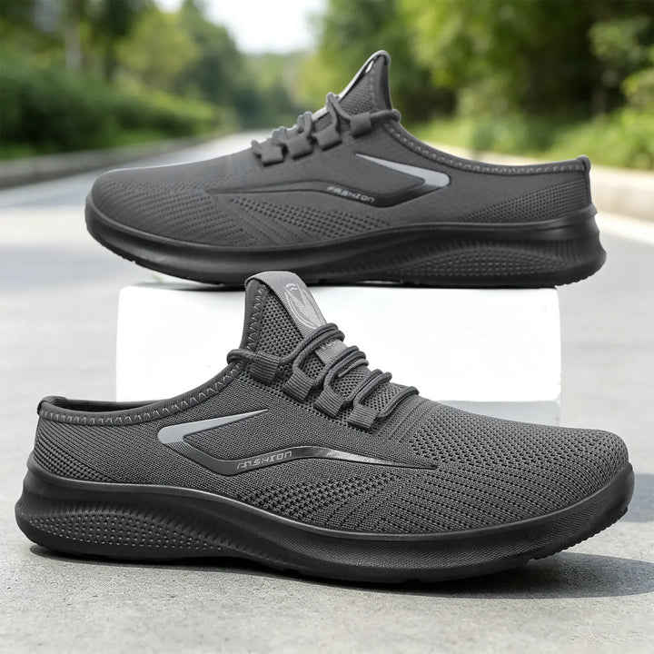A pair of dark grey Zylos breathable comfort slip-on trainers shown from a three-quarter angle, with a second pair slightly behind. The shoes feature engineered mesh uppers, elastic laces, and a matte black sole with a reflective side stripe.