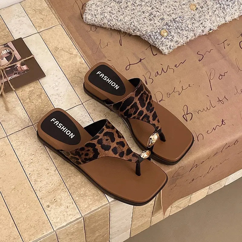 A pair of Zylos leopard print thong sandals with square toes and gold knot embellishments, resting on a textured surface with a cozy knit garment.