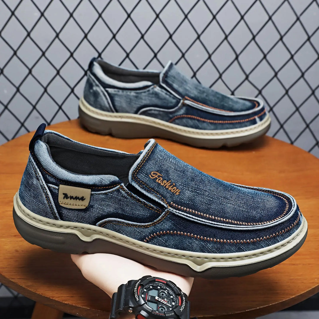 A pair of Zylos men's casual denim loafers in a blue wash, shown from a three-quarter angle, featuring orange contrast stitching, embroidered 'Fashion' detail, and a 'Tenne' label.