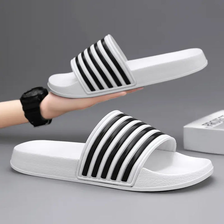 A pair of Zylos white and black striped EVA comfort slides, presented with one slide held by a hand against a studio grey background.