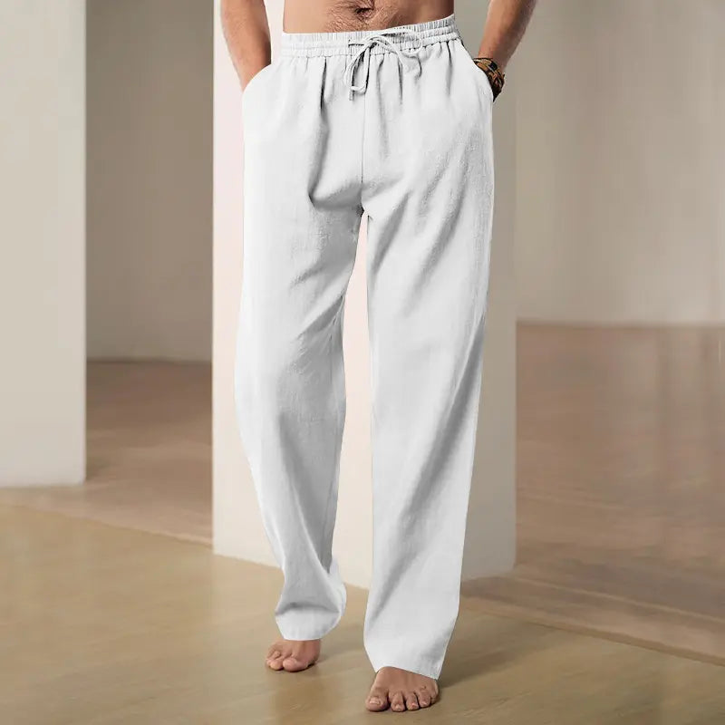 A man wears crisp white Zylos linen-blend wide-leg trousers with a drawstring waist and side pockets, styled casually in a relaxed interior setting.