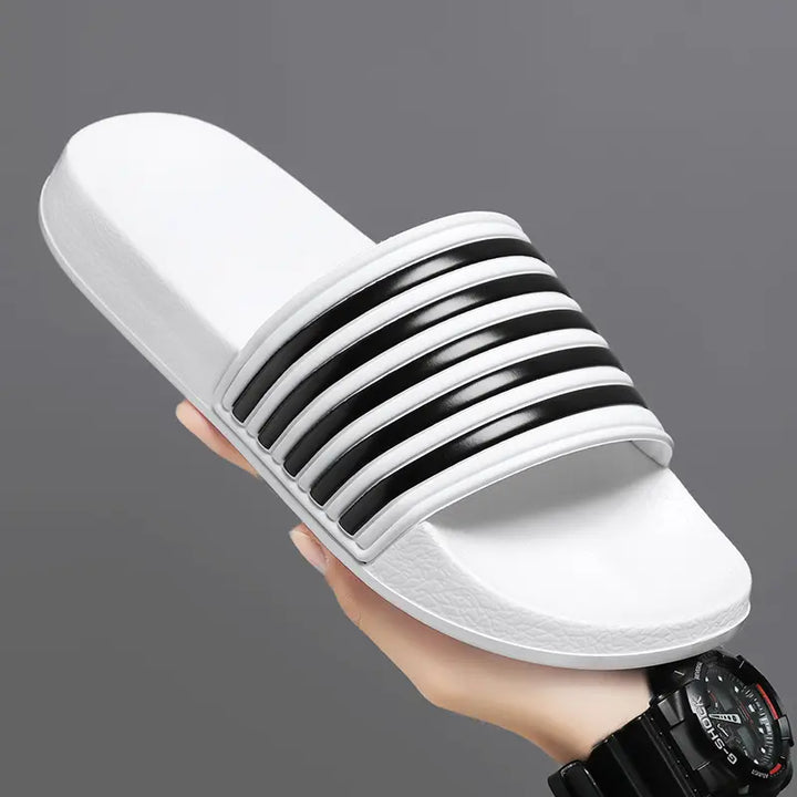 A white Zylos EVA Comfort slide with a black striped strap is held at a three-quarter angle, showcasing its thick sole and contoured footbed.