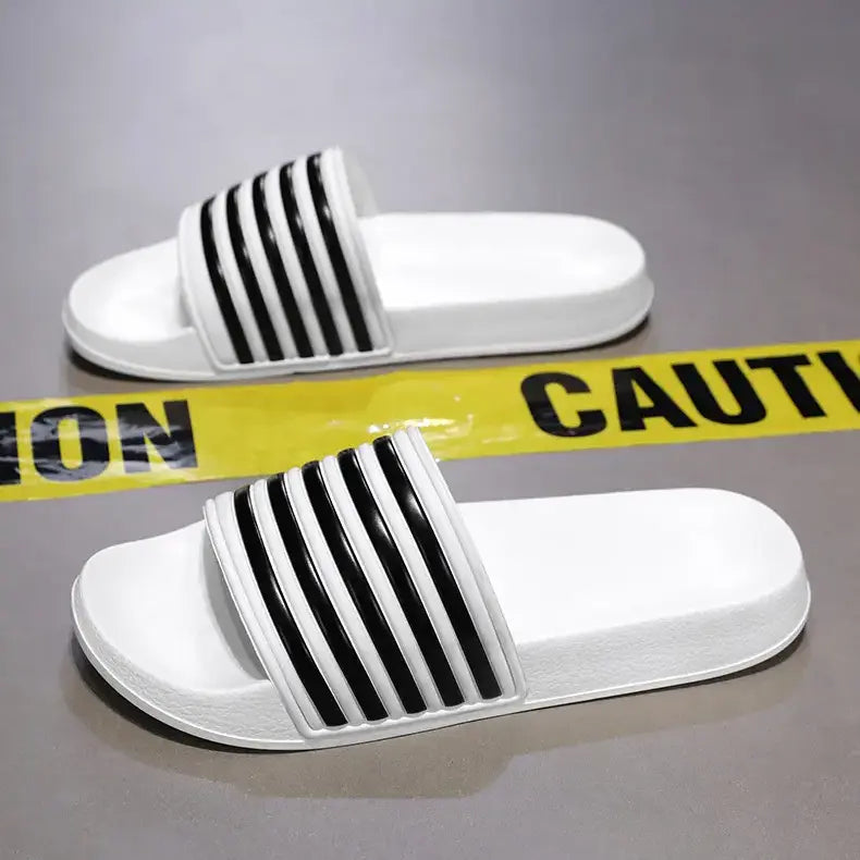 A pair of white Zylos EVA comfort slides featuring a wide strap with iconic black diagonal stripes, presented on a studio surface with yellow caution tape.