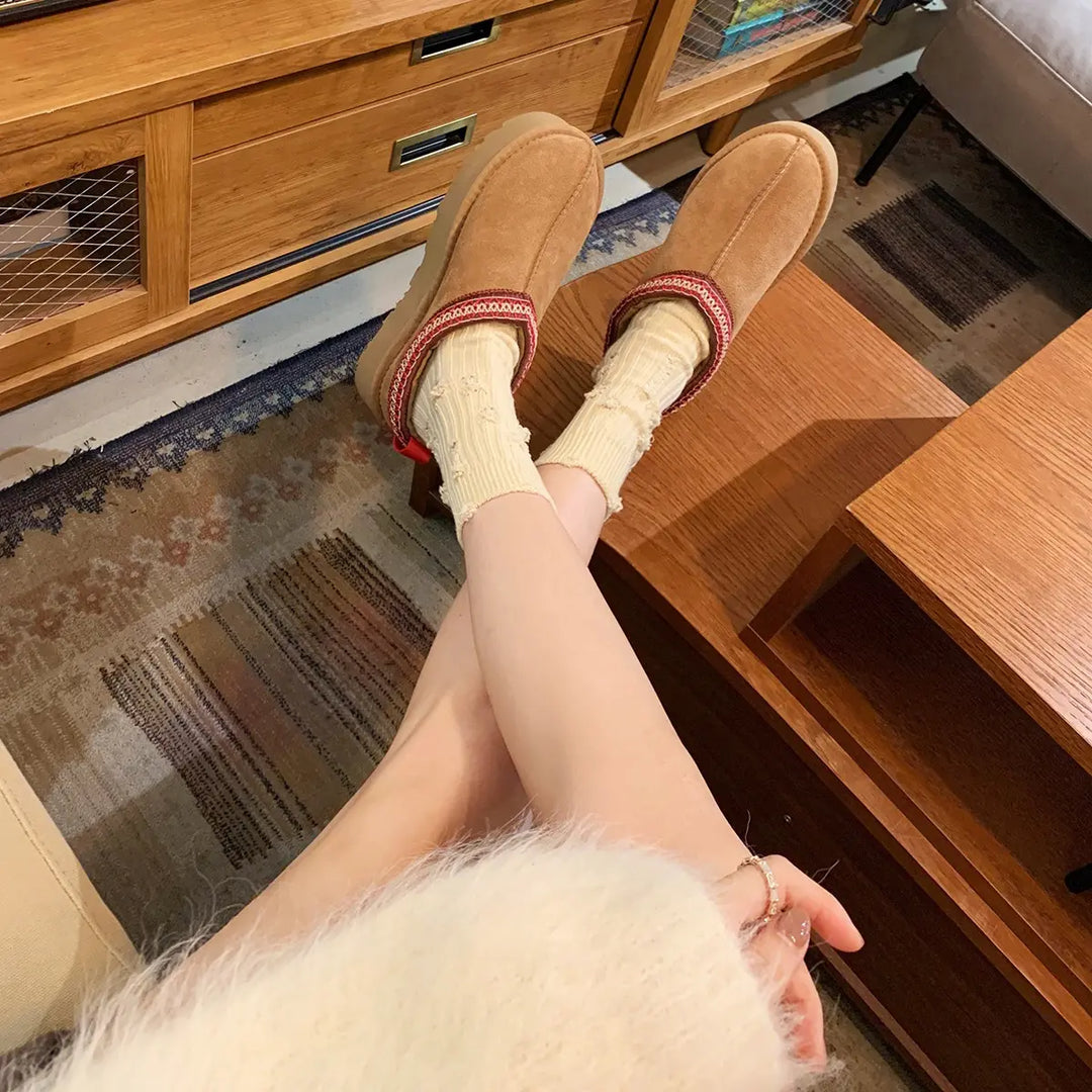 Cozy light tan suede-like women's low-cut boots with decorative red, white, and beige woven trim, worn with cream socks and a fluffy white sweater in a lifestyle setting.