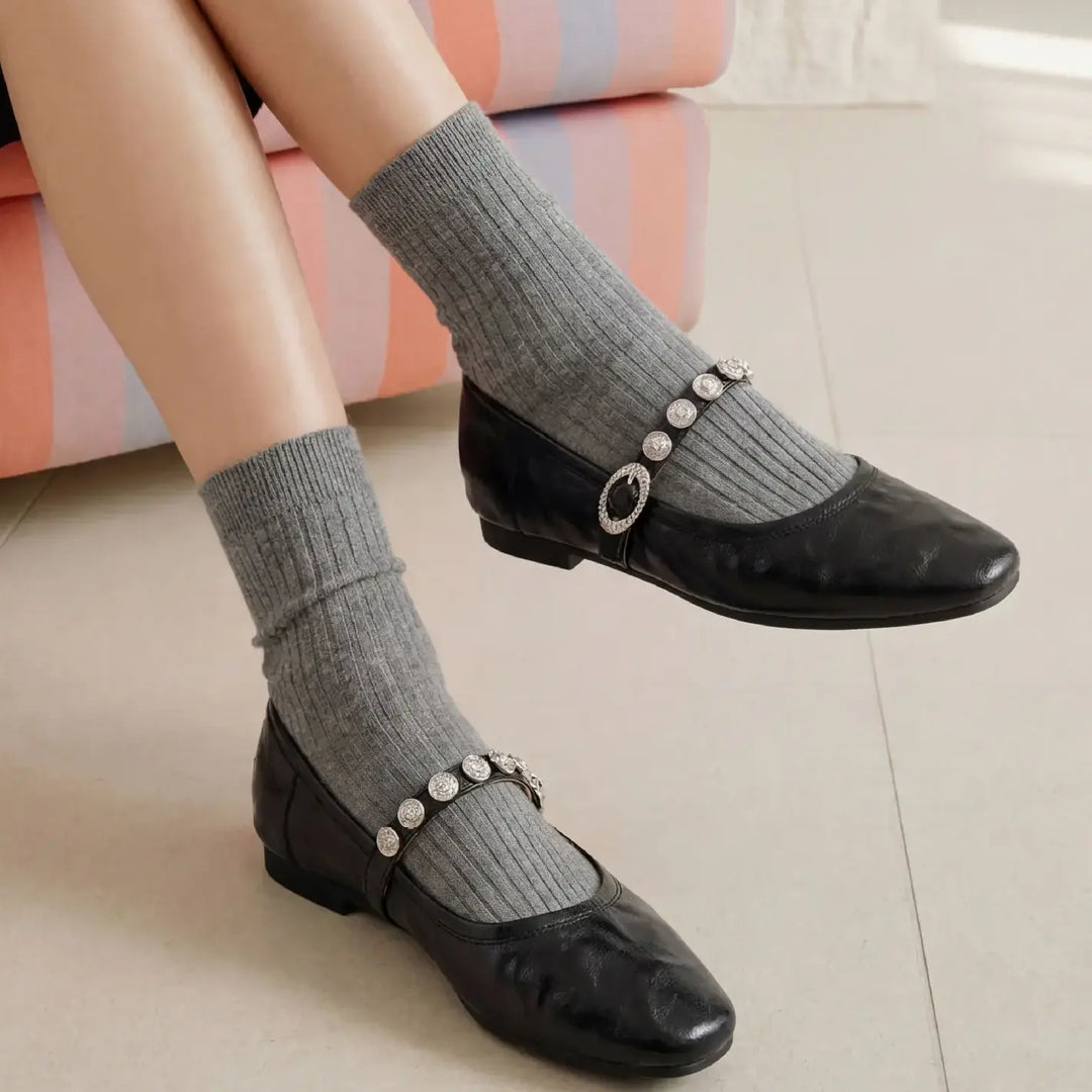 Black ruched leather Mary Jane flats with sparkling jeweled buckle straps, worn with grey socks by a seated model against a striped chair.