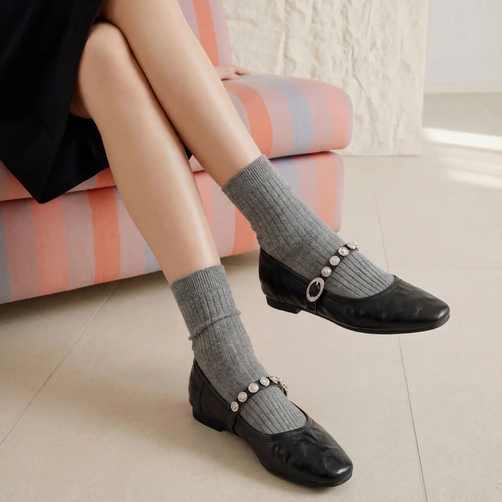 Black ruched leather Mary Jane flats with sparkling jeweled buckle straps, worn with grey socks by a seated model against a striped chair.