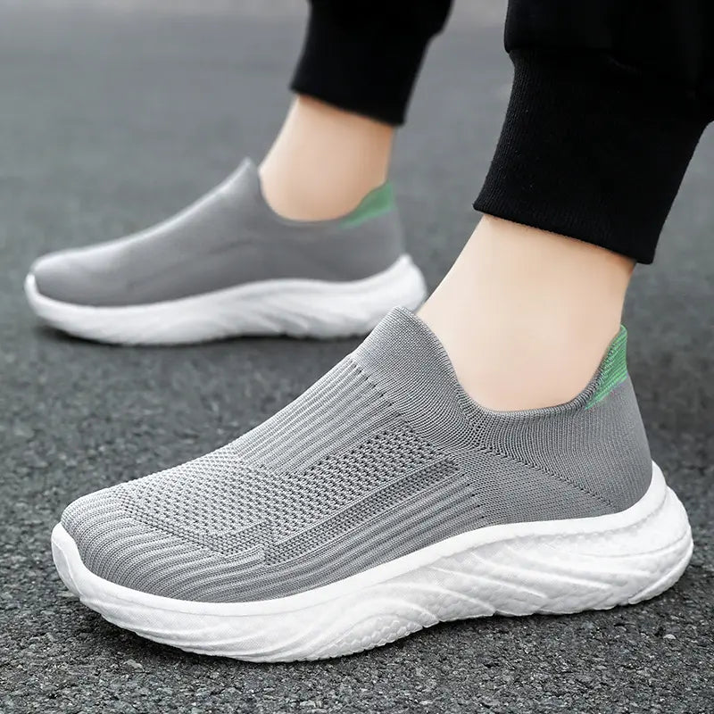 Zylpha men's casual grey slip-on sneakers made with breathable engineered knit, featuring a white sculpted sole and a subtle green heel accent, shown worn on asphalt.