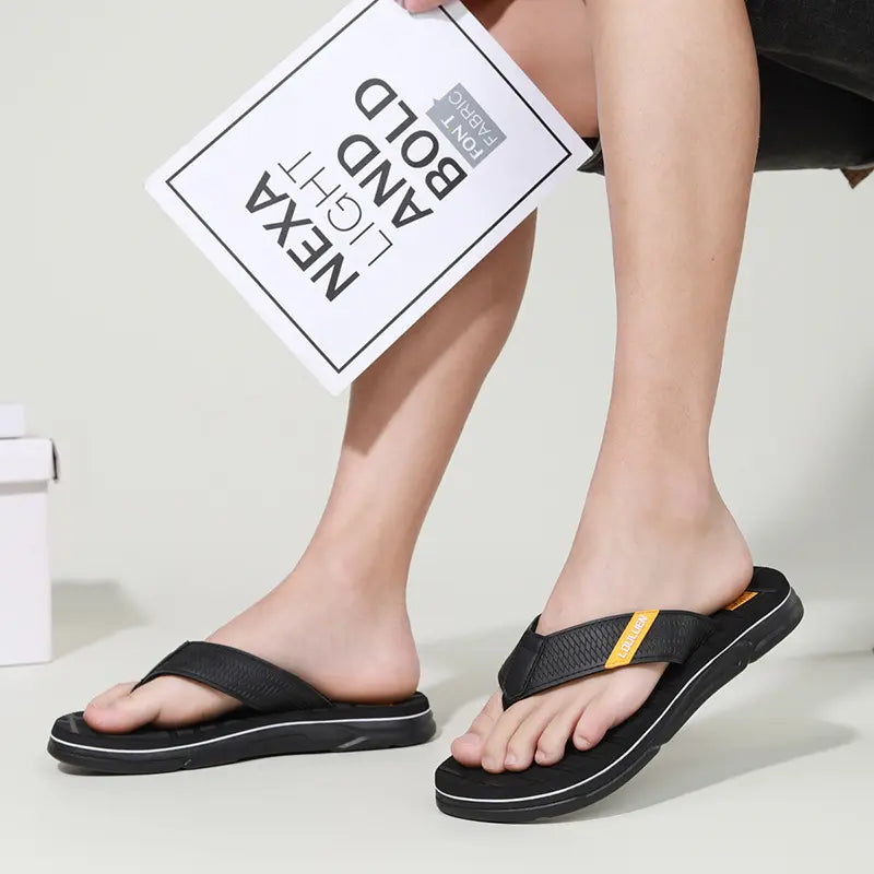 Men's black Zyndar flip-flops with textured crosshatch straps and a dark grey footbed with a grid pattern, worn by a person on a white studio background.