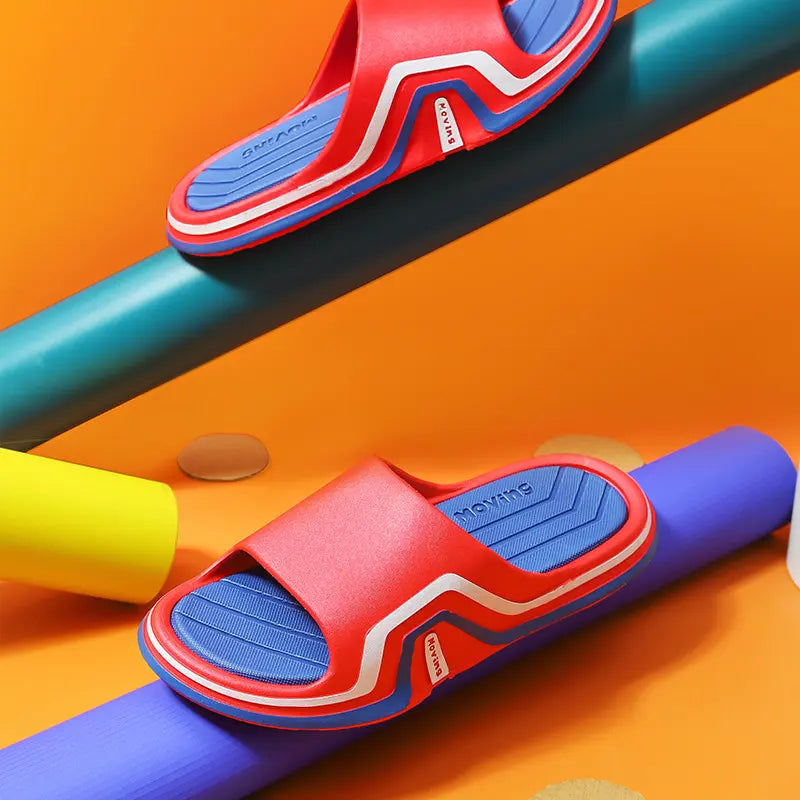 Red and blue unisex Zynex slides with white and red accent stripes on a layered EVA sole, presented in a vibrant studio setting with colored props. Features open toe and debossed 'MOVING' logo.