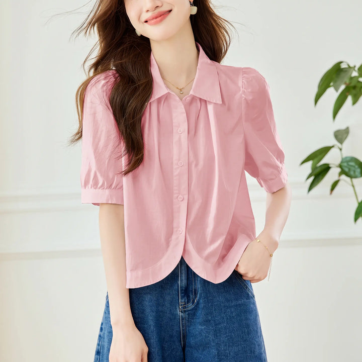 A woman models a soft pink short-sleeve blouse featuring puff sleeves, a pointed collar, button-down front, and a distinctive curved hemline, paired with dark denim jeans.