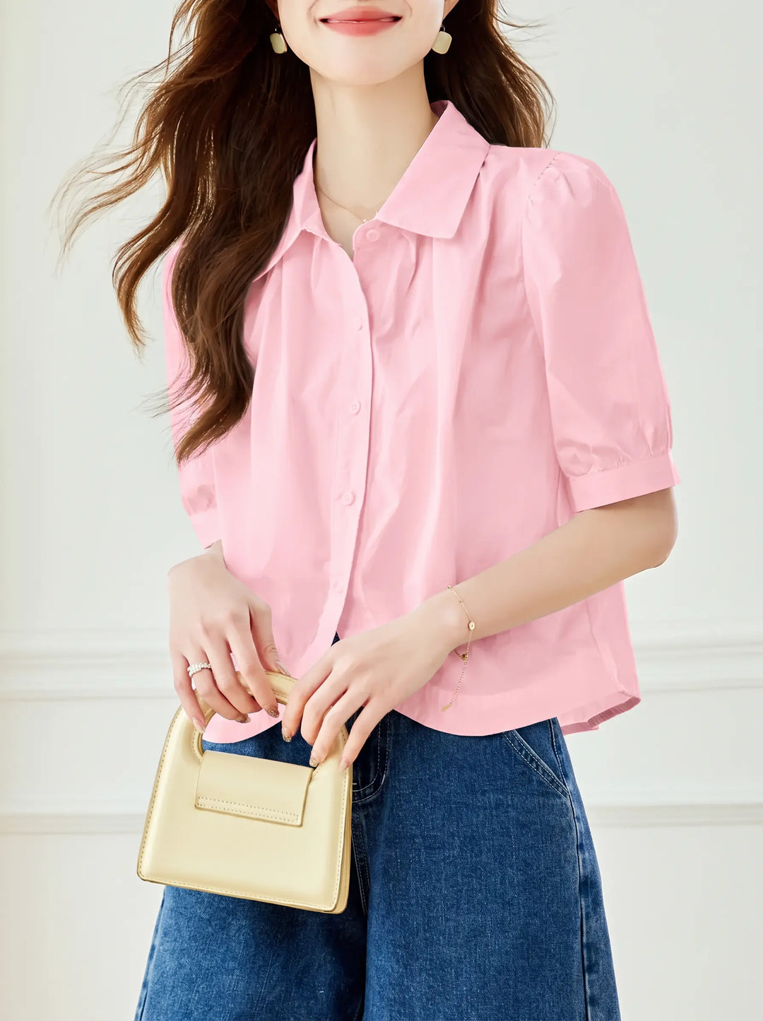 A woman models a Soft Petal Pink short-sleeve blouse featuring a classic pointed collar, subtle puff sleeves, and a clean button-down front, paired with denim shorts.