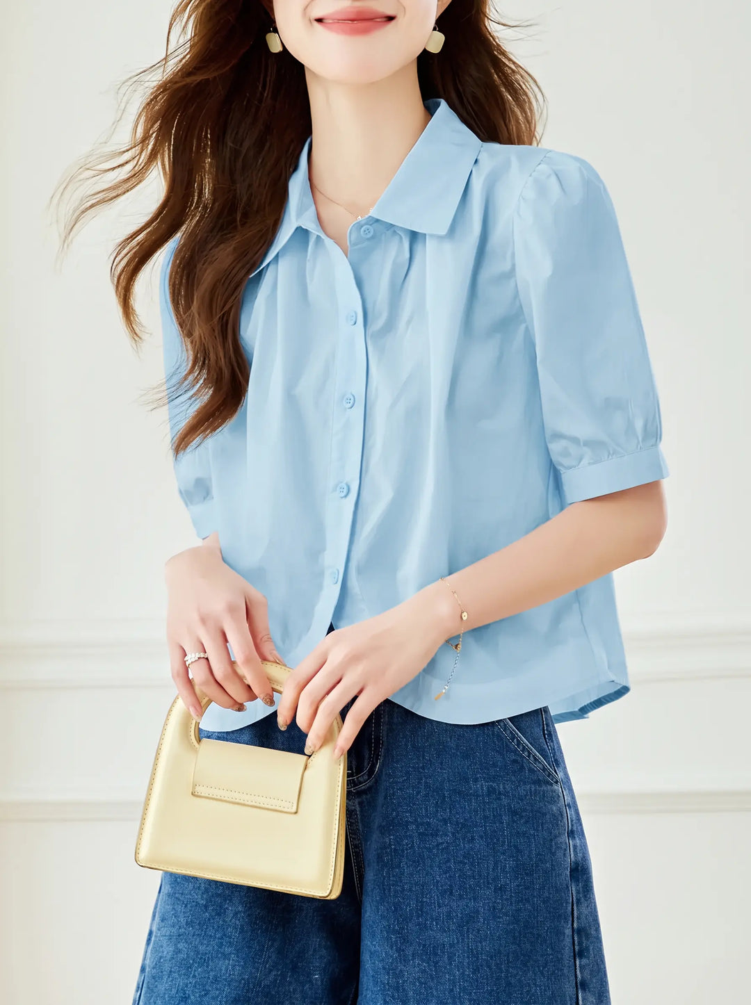 A woman wears a powder blue Zyphia short-sleeve blouse with puff sleeves and a curved hem, paired with denim and holding a gold handbag.