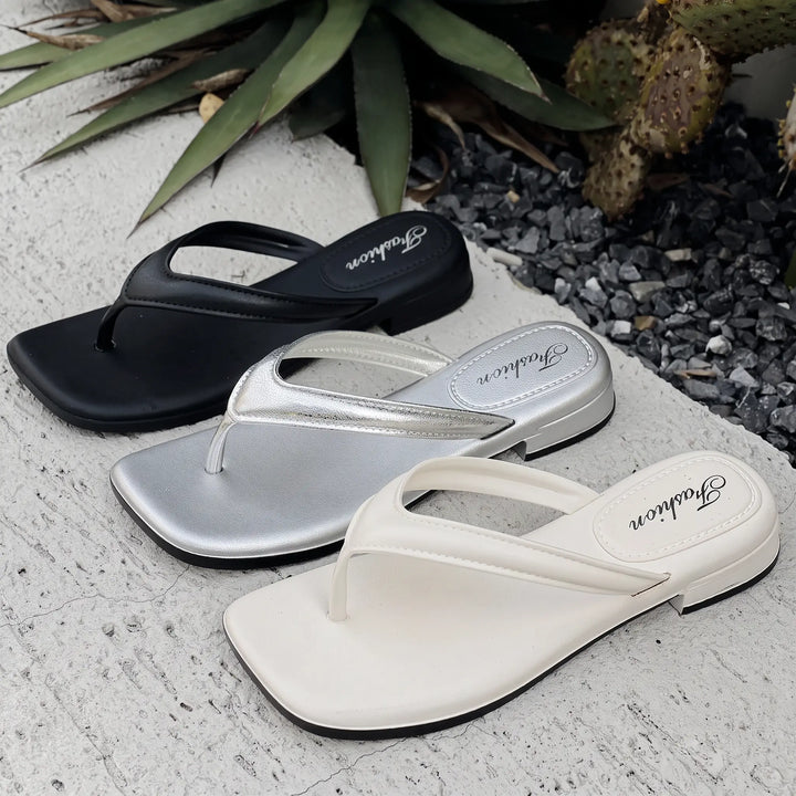 Three pairs of Zyphyr chic square-toe thong sandals displayed in black, shimmering silver, and pure white, set against a textured outdoor backdrop with lush greenery.