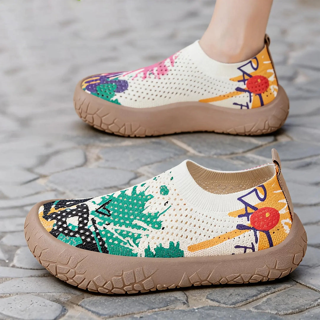 Zyra artistic print slip-on flat shoes with a breathable knit upper and a taupe rubber sole, shown on a foot in an outdoor setting.