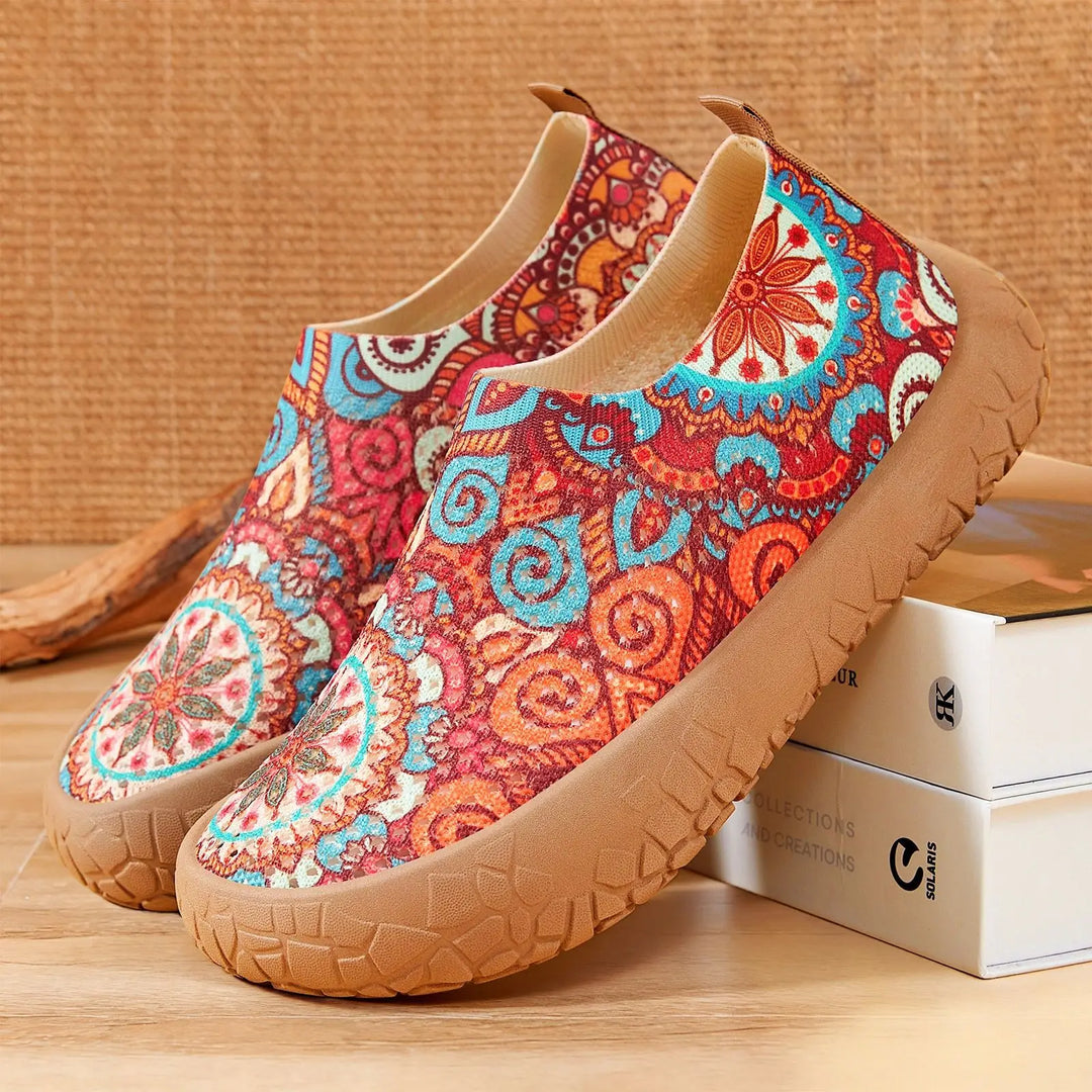Zyra artistic print slip-on flats in bohemian design with vibrant orange, red, and blue patterns, showcasing a comfortable knit upper and taupe rubber sole from a three-quarter angle.