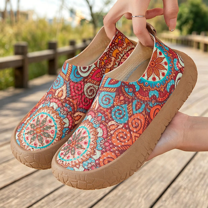 Zyra artistic print slip-on flats with a vibrant bohemian pattern in red, orange, and blue, shown held outdoors on a wooden pathway.