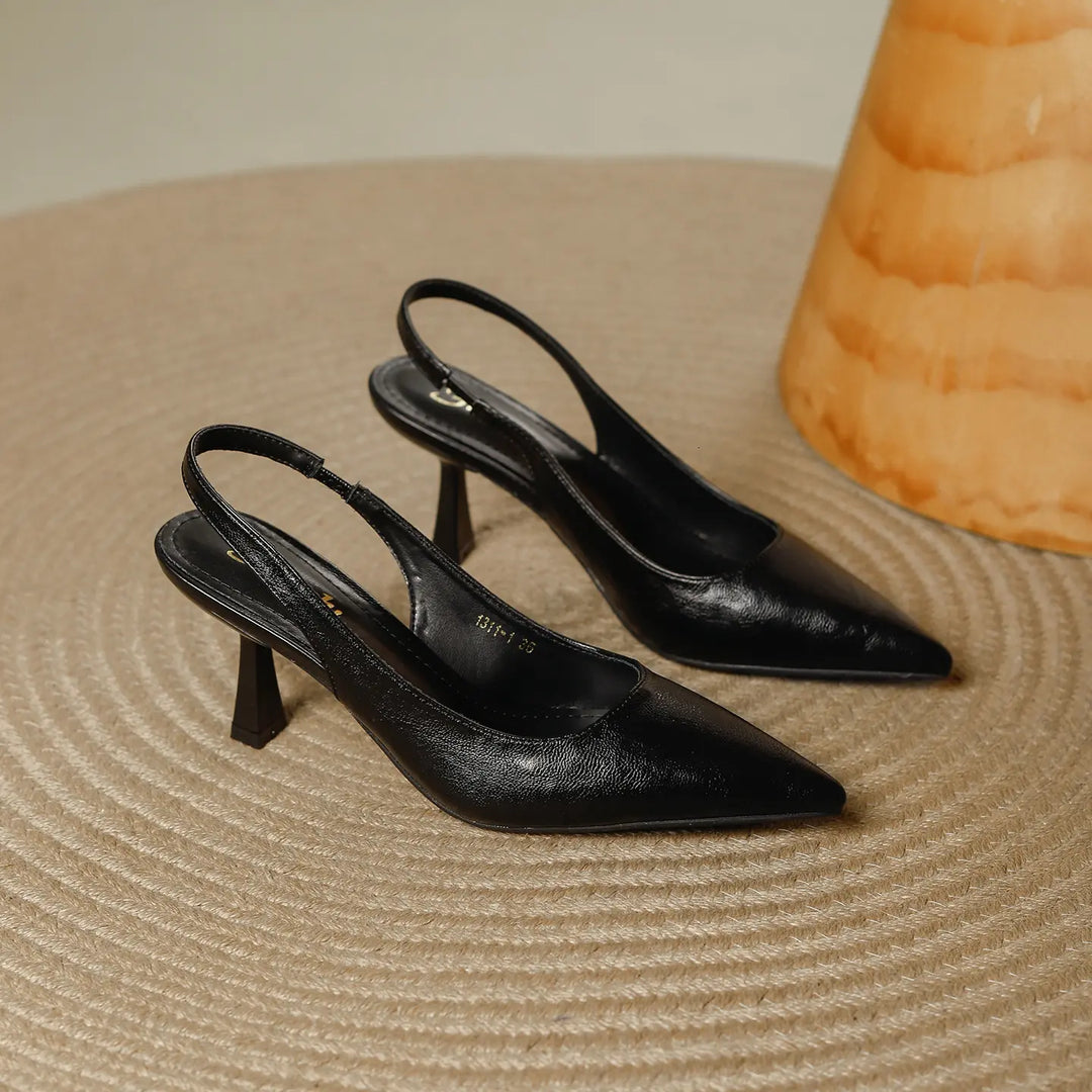 Black pointed toe slingback heels with a sculpted stiletto heel and elasticated strap, presented on a textured surface with a decorative vase in the background.