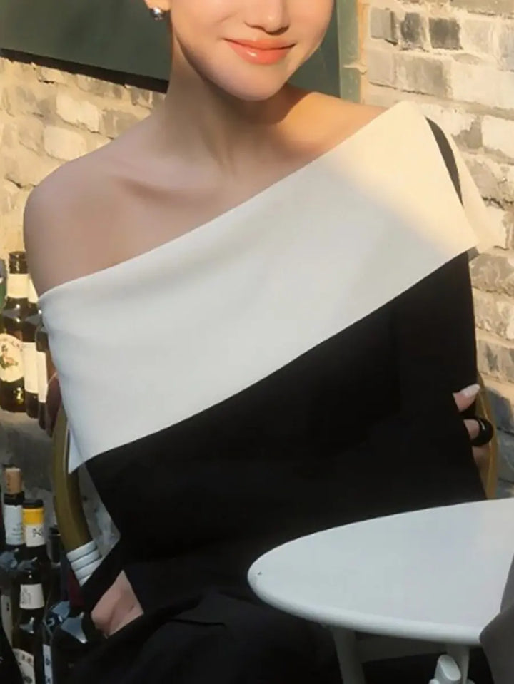A Zythos asymmetrical knit top in black and optic white, worn by a person seated, showcasing the striking off-shoulder neckline and color-block design. The soft fabric drapes elegantly in a lifestyle setting.