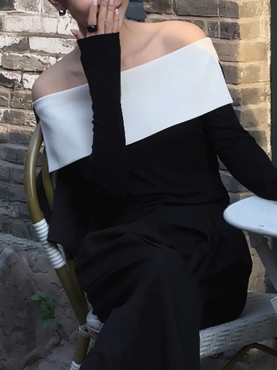 Woman wearing a black and optic white asymmetrical off-shoulder knit top, seated outdoors with a brick wall in the background.