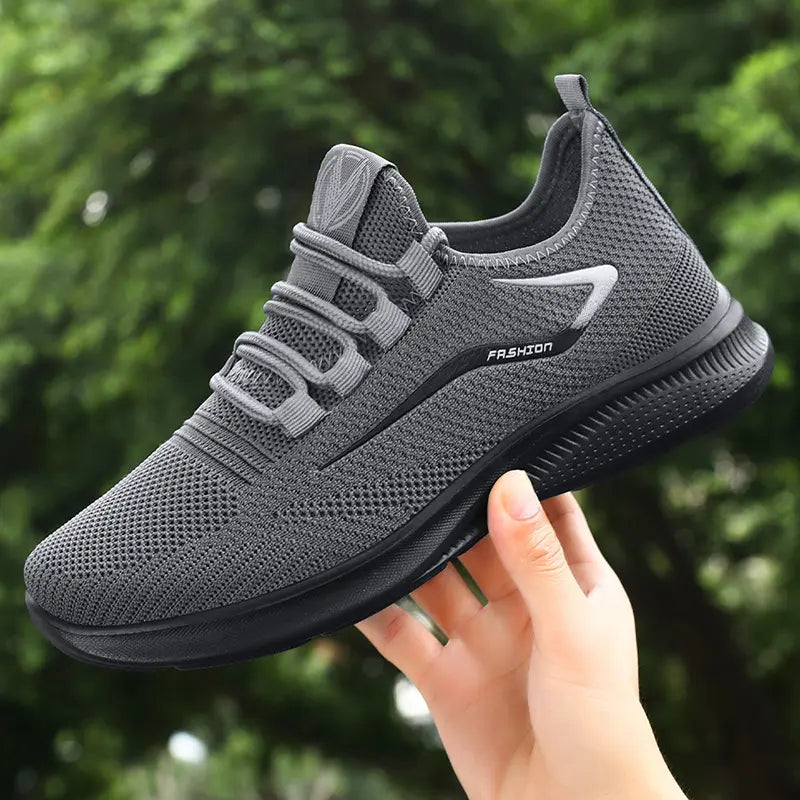 A grey Zythos men's breathable trainer held by a hand, showcasing its engineered knit upper and matte black sole against a blurred outdoor greenery.